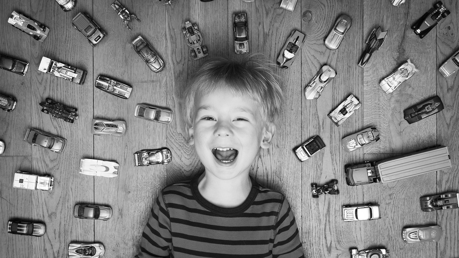 En dreng ligger på gulvet omgivet af legetøjsbiler og smiler stort
