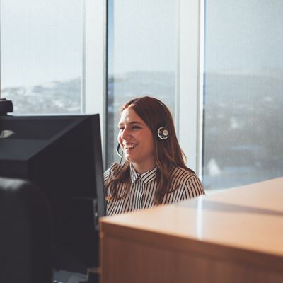 Kvinde som sidder i et kontor forand en computer med headset på. 