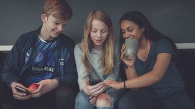 Tre teenagere sidder på en bænk og kigger i en mobil. 